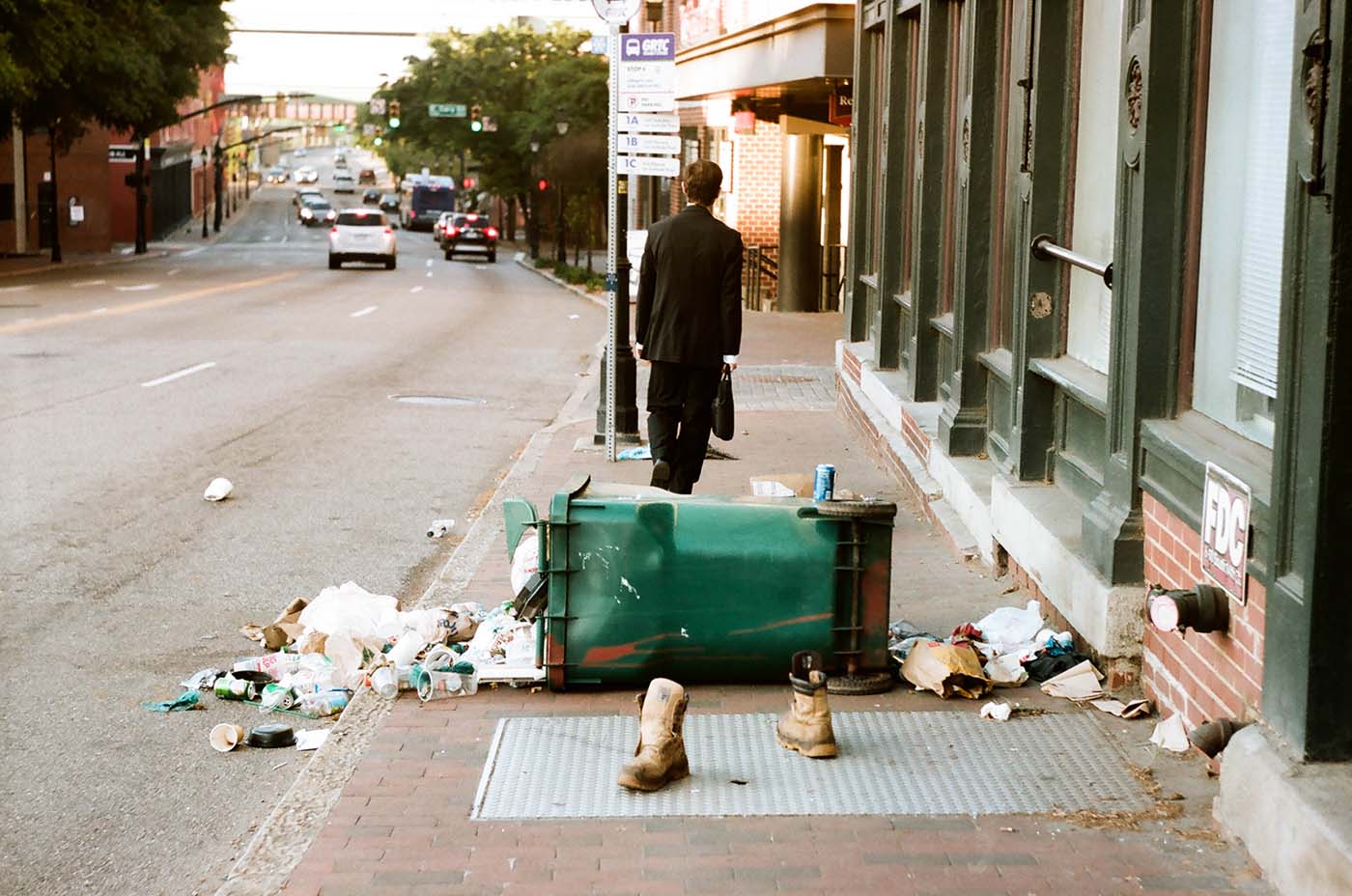 Trash can turned over with garbage spilt, boots standing nearby and man wearing a suit walking away.